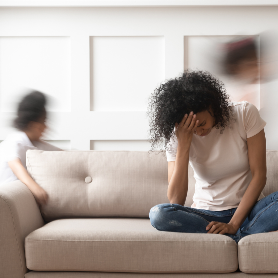 Person sitting on a couch with their head in their hand while children move around in the background, conveying feelings of stress and overwhelm.